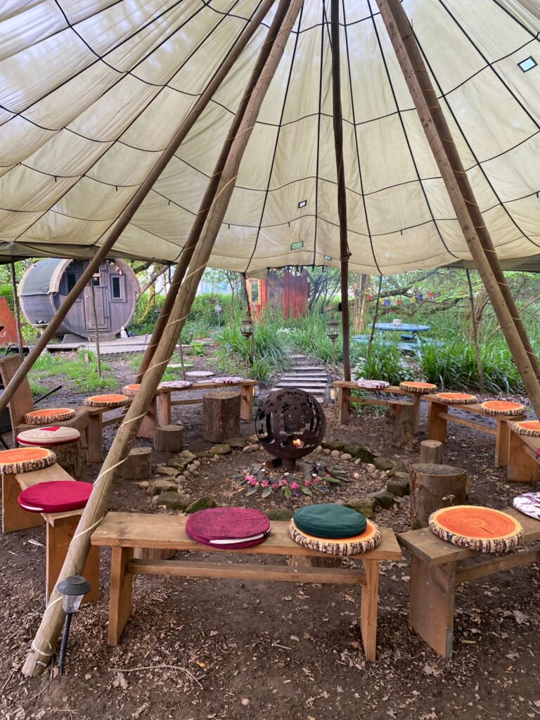Photo of the Circle space. Fire in the centre with benches and orange cushions around. Nature surrounds the space and there is a tarpaulin tent roof,