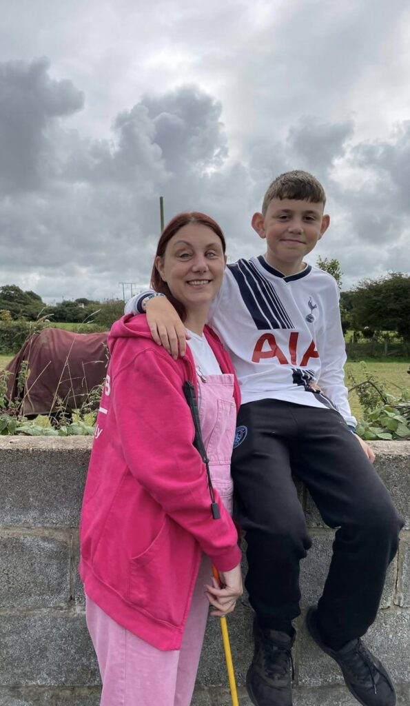 Holiday photo of Nina and son Dylan. Dylan is sitting on a wall with his arm around Nina's shoulder. Background of countryside and horses.